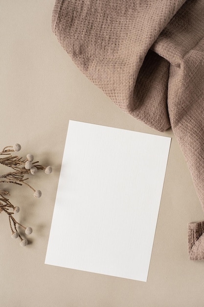 Blank paper sheet card with mockup copy space and dry floral branch and blanket cloth on neutral beige background Minimal aesthetic wedding invitation template Flat lay top view