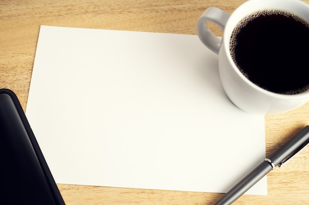 Blank paper on wooden desk table with cup of coffee, pen and smartphone. Mock up template. Study or business concept