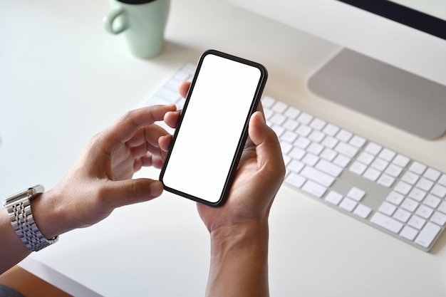 Blank screen mobile phone in male's hand on studio workspace with office supplies.