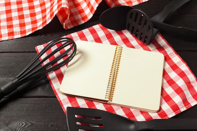 Blank sheet of opened notepad and kitchen utensils on  table with tablecloth, copy space