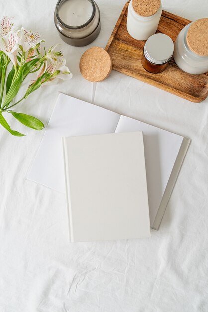 Blank white book, coffee, glass jars and flowers on white bed, flat lay