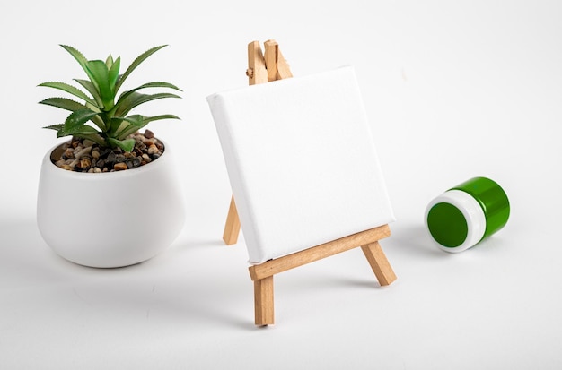 Blank white canvas mockup in perspective angle mini wood tripod easel and potted house plant green paint in jar on desk