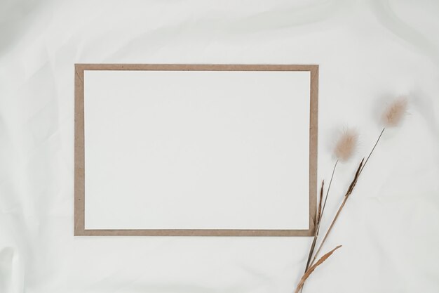 Blank white paper on brown paper envelope with Rabbit tail dry flower on white cloth. horizontal blank greeting card. Top view of Craft envelope on white background