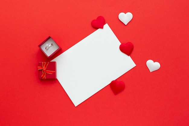 blank white paper on red background with hearts and ring in gift box. Valentine's day background. Mockup of love letter. Flatly, top view.