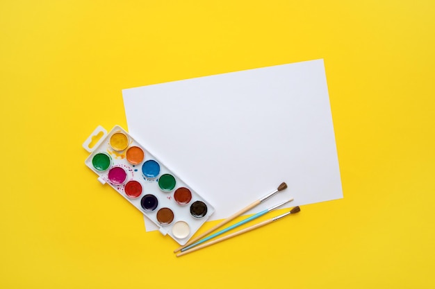 Blank white sheet of paper with watercolors and brushes on a yellow background