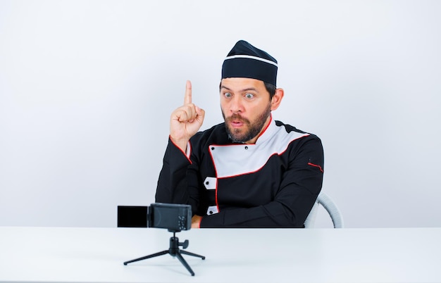 Blogger chef is pointing up with forefinger by sitting in front of his mini camera on white background