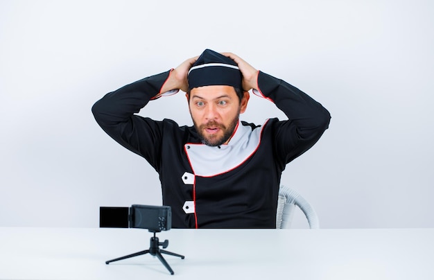 Blogger chef is posing to his mini camera by putting hands behind head on white background