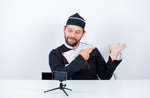 Blogger chef is raising up his hand and pointing it with forefinger by sitting in front of his mini camera on white background