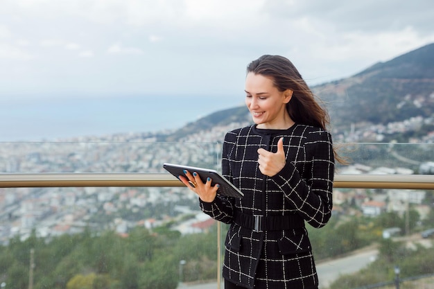 Blogger girl is holding planshet computer by showing perfect gesture against the background of city view