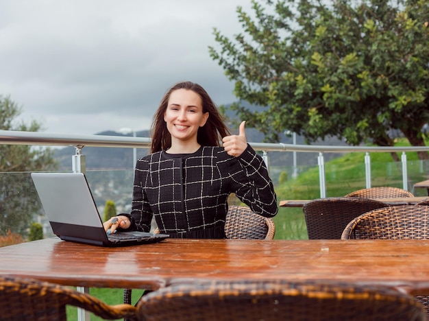 Blogger girl with laptop is showing perfect gesture to camera by sitting on nature view background