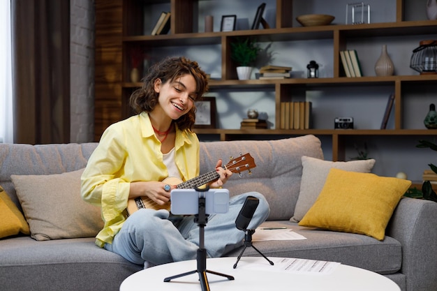 Blogger with mobile phone and microphone A beautiful hipster girl in a yellow shirt plays and sings on the ukulele on the sofa in the apartment Online learning to play the guitar