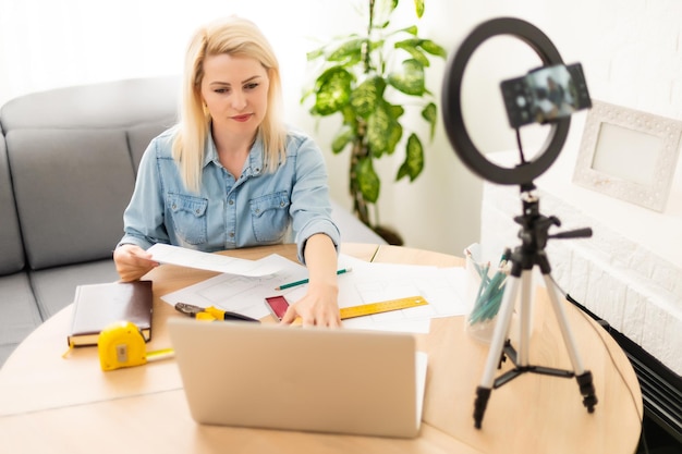 blogger woman recording video on smartphone and lighting herself with ring lamp at table in light room.