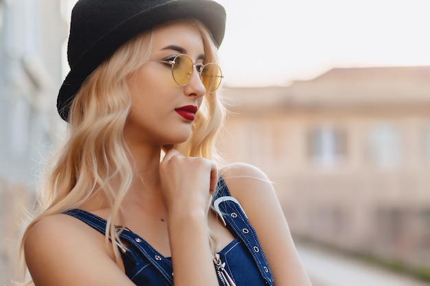 Blond attractive girl in sunglasses and stylish hat at summer sunshine urban