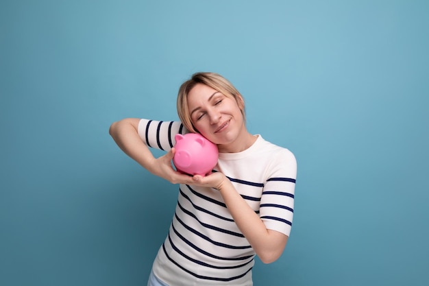 Blond attractive young woman in casual attire carefully hugging a piggy bank with savings on a blue