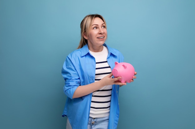 Blond attractive young woman in casual attire with a smile holds a piggy bank with savings in her