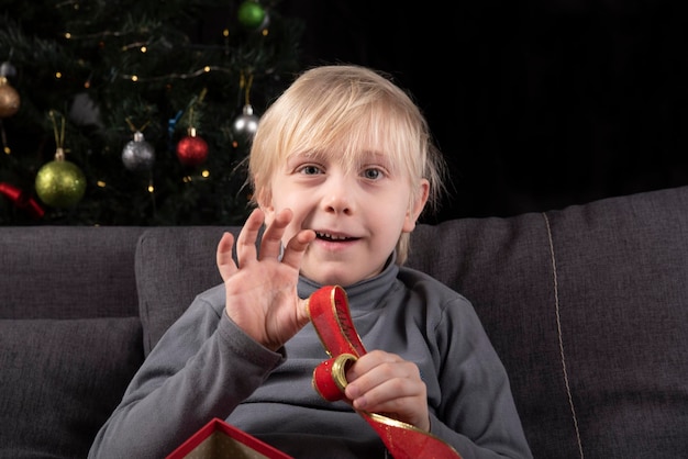 Blond boy holding Christmas tinsel in his hands and waving at the camera sitting on the sofa against the background of the Christmas tree.