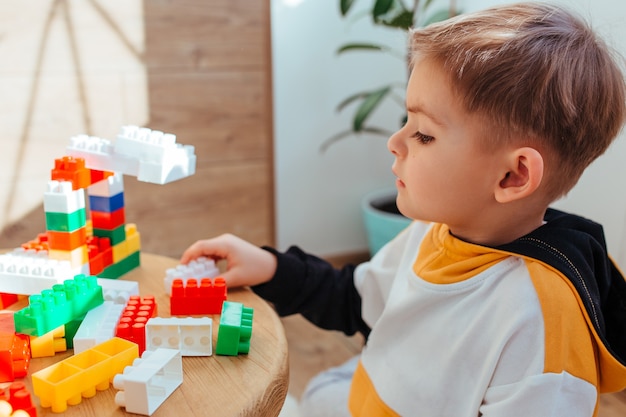 A blond boy is playing with a construction kit, with a wooden wall in the background. close-up