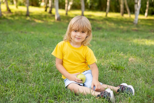 a blond boy is sitting on the lawn in a yellow T-shirt holding a green apple in his hands