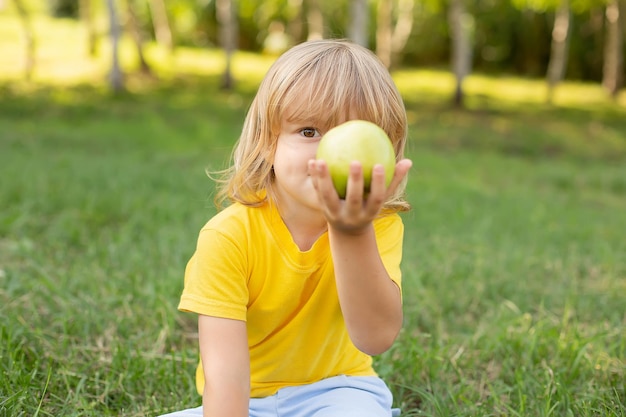 a blond boy is sitting on the lawn in a yellow T-shirt holding a green apple in his hands