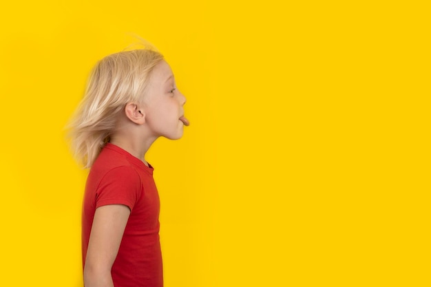 Blond boy in red shirt shows tongue Portrait side view on yellow background Copy space