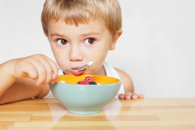 A blond boy with brown eyes in a white T-shirt eats cottage cheese with berries at a wooden table.