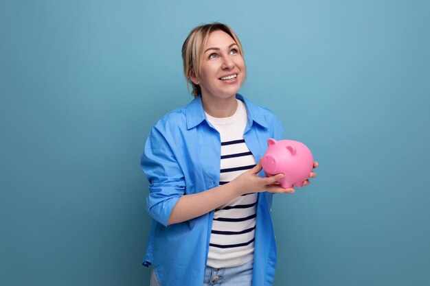 Blond cheerful girl in casual attire carefully holds a piggy bank with savings on a blue background