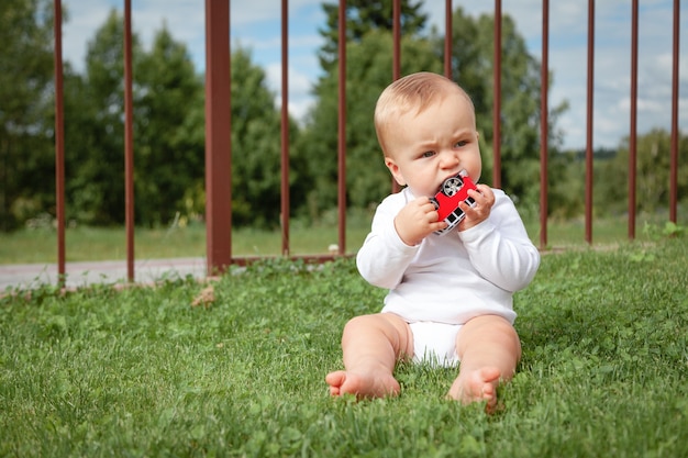 Blond cute little blue-eyed baby in white bodysuit sitting in green grass and playing with car toy.