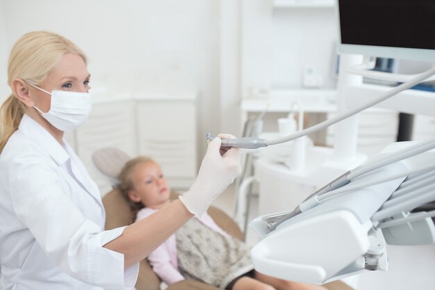 A blond doctor holding a dental drill while working with her little patient