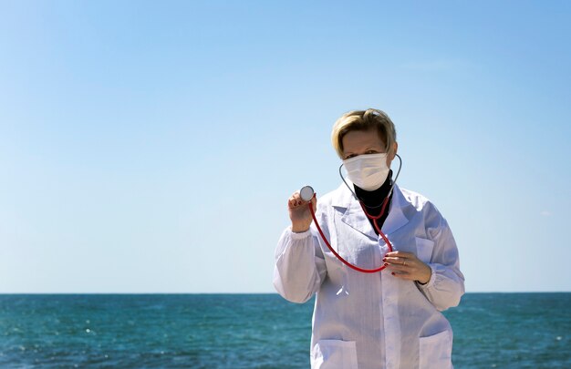 Blond doctor woman of Russian Caucasian appearance Holding A Stethoscope In Hand. Portrait of professional confident American doctor in medical mask and white coat, stethoscope over neck on a beach