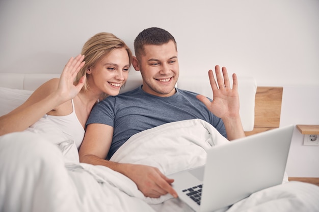  blond female and handsome brunette male staying in bedroom while talking online on laptop and waving