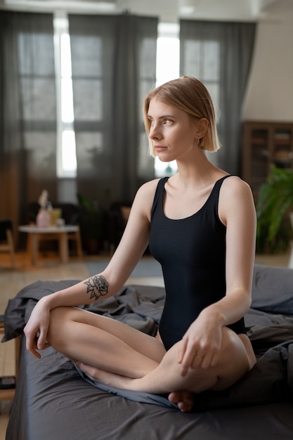 Blond girl in black leotard crossing legs while sitting on bed against window and bedroom interior and thinking of plans for new day