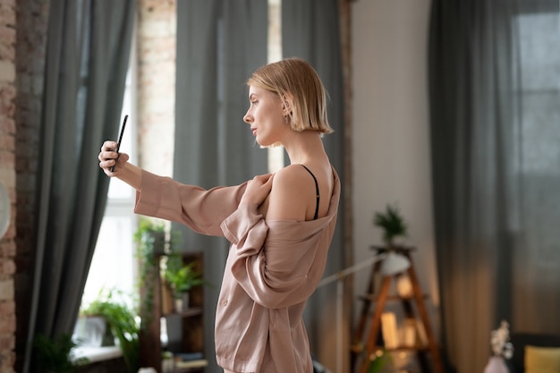 Blond girl in black lingerie and beige pajamas standing in front of large window in bedroom and making selfie or recording video for blog