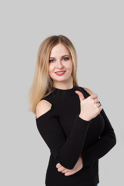 Blond girl is making a thumbs up sign and smiling. the background is light gray.