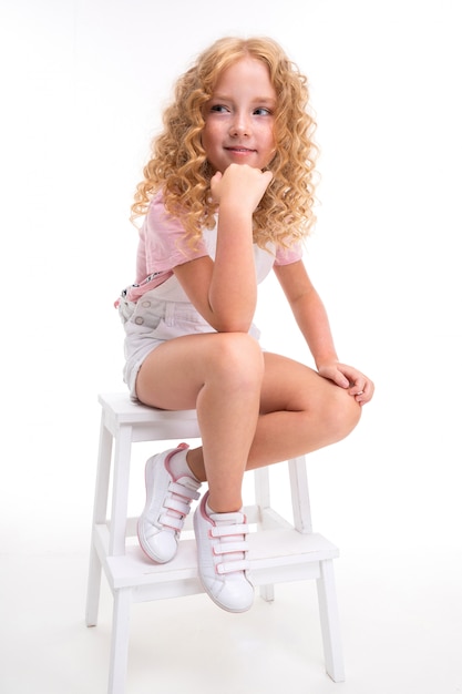 Blond girl sitting on chair and posing on white wall