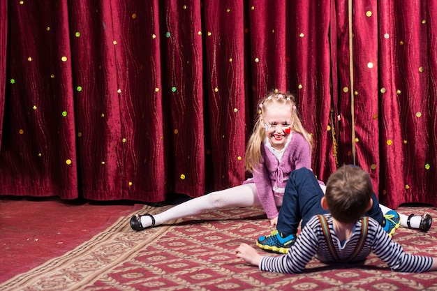 Blond Girl Wearing Clown Make Up Performing Splits on Stage with Red Curtain with Young Boy in Foreground