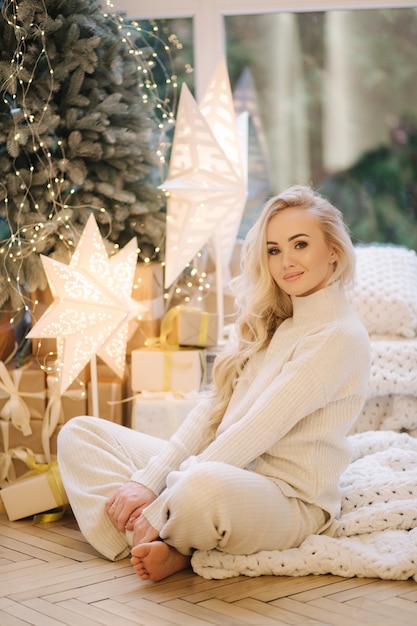 Blond hair woman in white knitted suit sitting at home by Christmas tree. Beautiful woman at winter