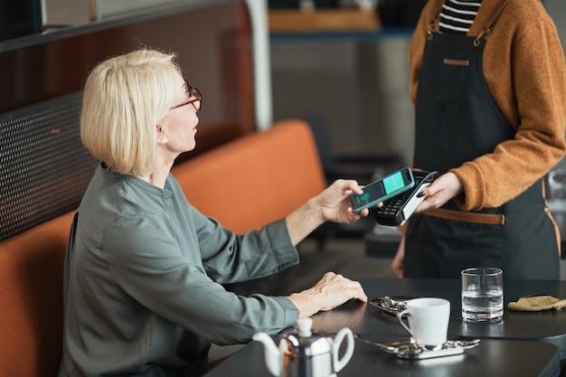 Blond-haired senior Caucasian woman sitting at table in coffee shop and putting smartphone to payment terminal held by waitress in apron