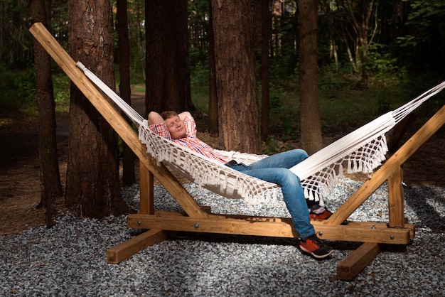 Blond man in jeans and shirt is resting in hammock outdoors in backyard