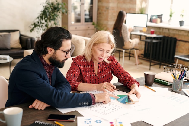 Blond mature female designer and her bearded male colleague pointing at color palette while working over new creative project in office