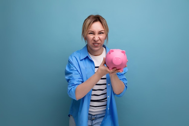 Blond neat cheerful girl with a grimace holds a piggy bank on a blue background