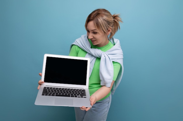 Blond positive cute female freelancer in casual outfit showing screen with mockup on laptop on blue