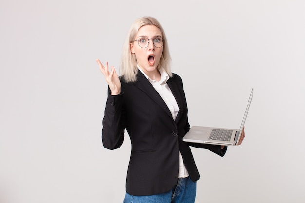 Blond pretty woman feeling extremely shocked and surprised and holding a laptop