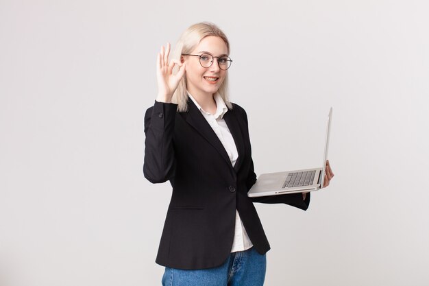 Blond pretty woman feeling happy, showing approval with okay gesture and holding a laptop