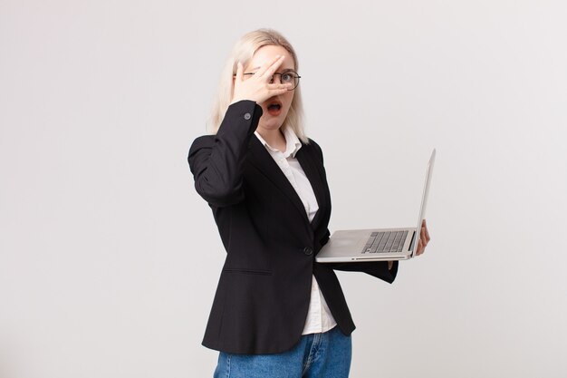 Blond pretty woman looking shocked, scared or terrified, covering face with hand and holding a laptop