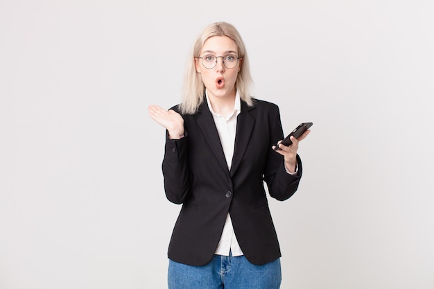 blond pretty woman looking surprised and shocked, with jaw dropped holding an object and holding a mobile telephone