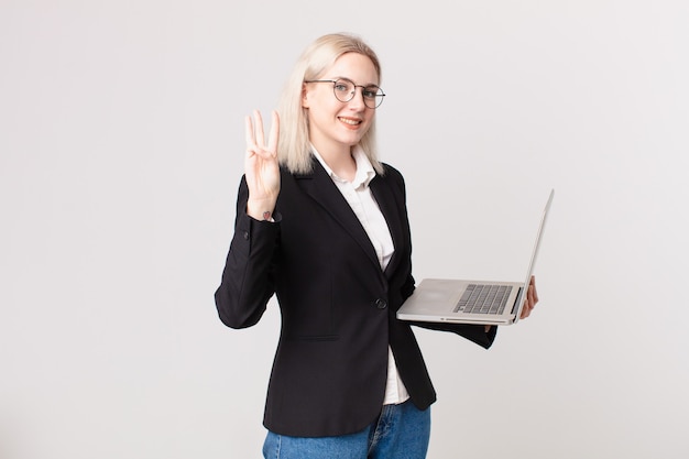 Blond pretty woman smiling and looking friendly, showing number three and holding a laptop