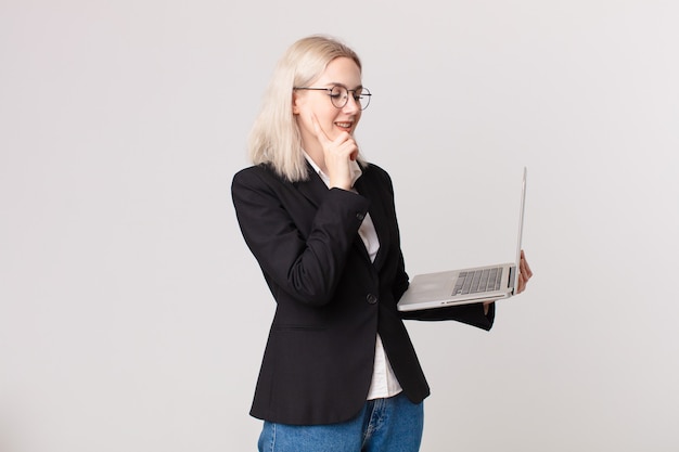 Blond pretty woman smiling with a happy, confident expression with hand on chin and holding a laptop