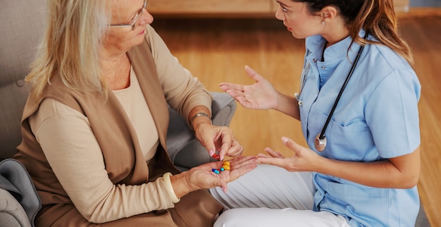 Blond senior woman sitting at home and holding hand full of pills. Nurse sitting next to her and giving her and advice.