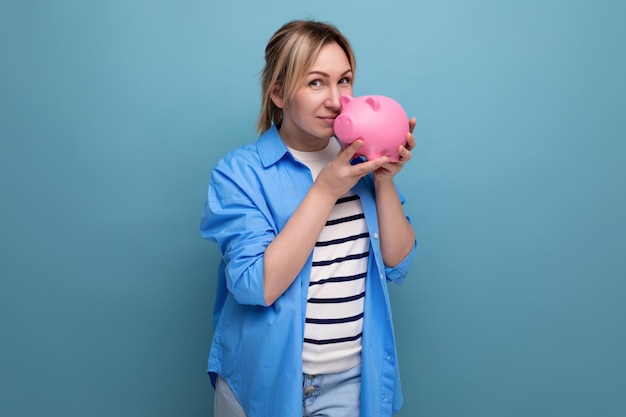 Blond smiling girl in casual outfit holding a piggy bank on a blue background with copy space