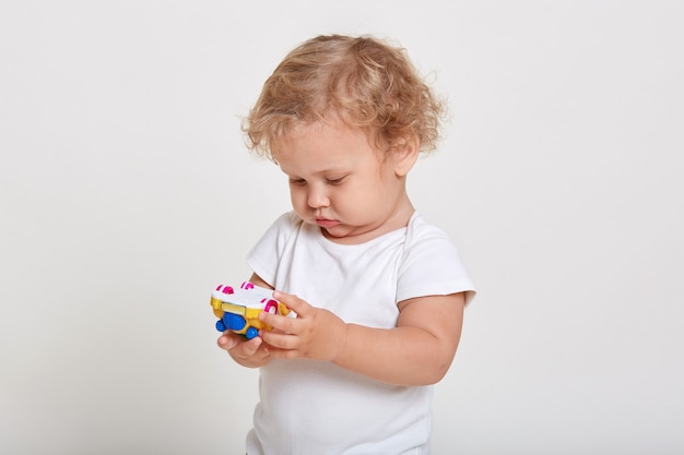 Blond toddlers playing with color car isolated on white space, looking concentrated at toy in his hands